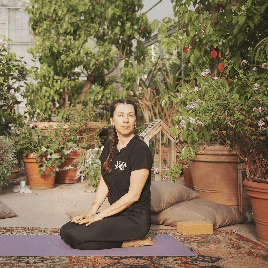 Sara Serroni, insegnante di Yoga Vinyasa, seduta in meditazione su un tappetino in un giardino con piante